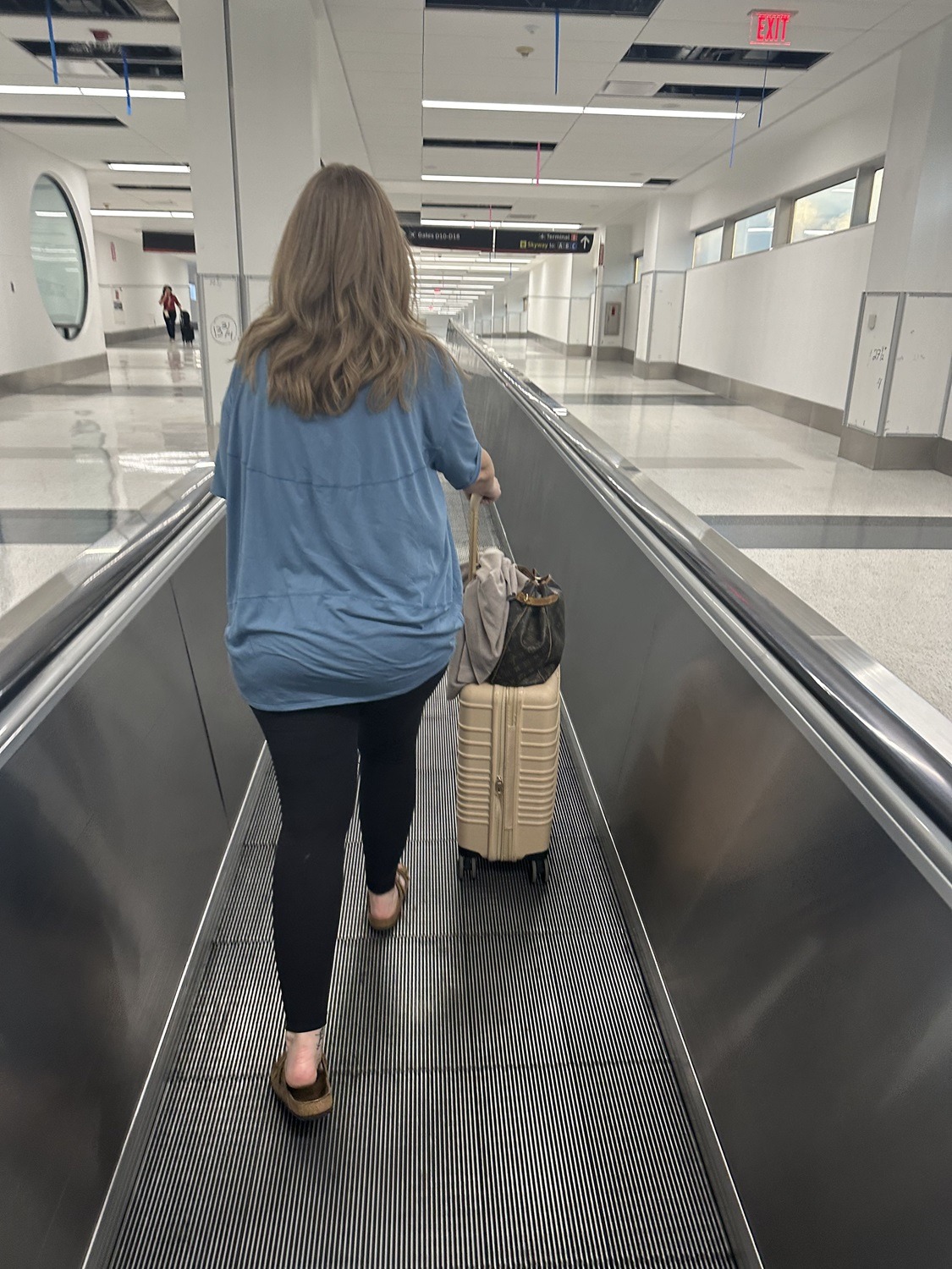 Brandie moving away on an airport walkway, wheeling her carryon; in her happy place on another trip she didn't have to plan